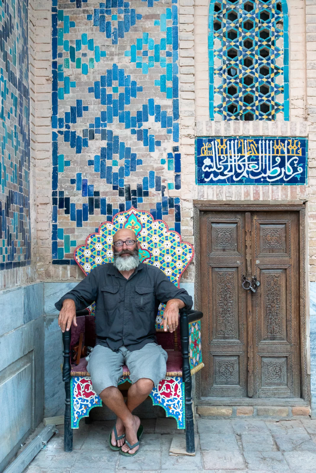 Franse fietser met grijze baard op een beschilderde stoel bij een betegelde deur in Samarkand