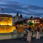 Verlichte ronde vestingtoren met Turkse vlag aan de rand van de oude stad van Antalya bij avond, met mensen op de muur en verlichte terrassen erachter