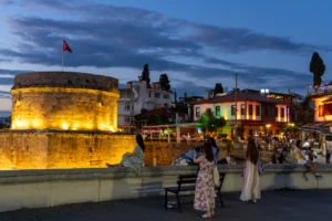Verlichte ronde vestingtoren met Turkse vlag aan de rand van de oude stad van Antalya bij avond, met mensen op de muur en verlichte terrassen erachter