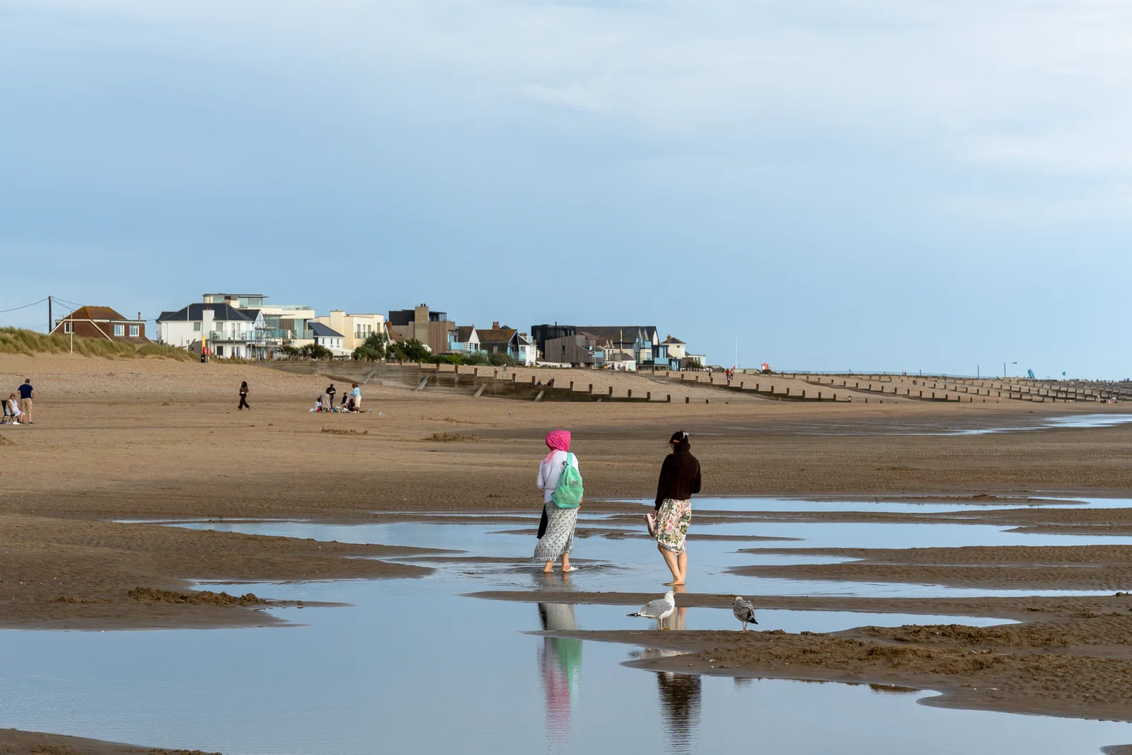 Twee wandelaars op het brede natte strand van Camber Sands bij eb, met vakantiehuizen en een houten golfbreker op de achtergrond