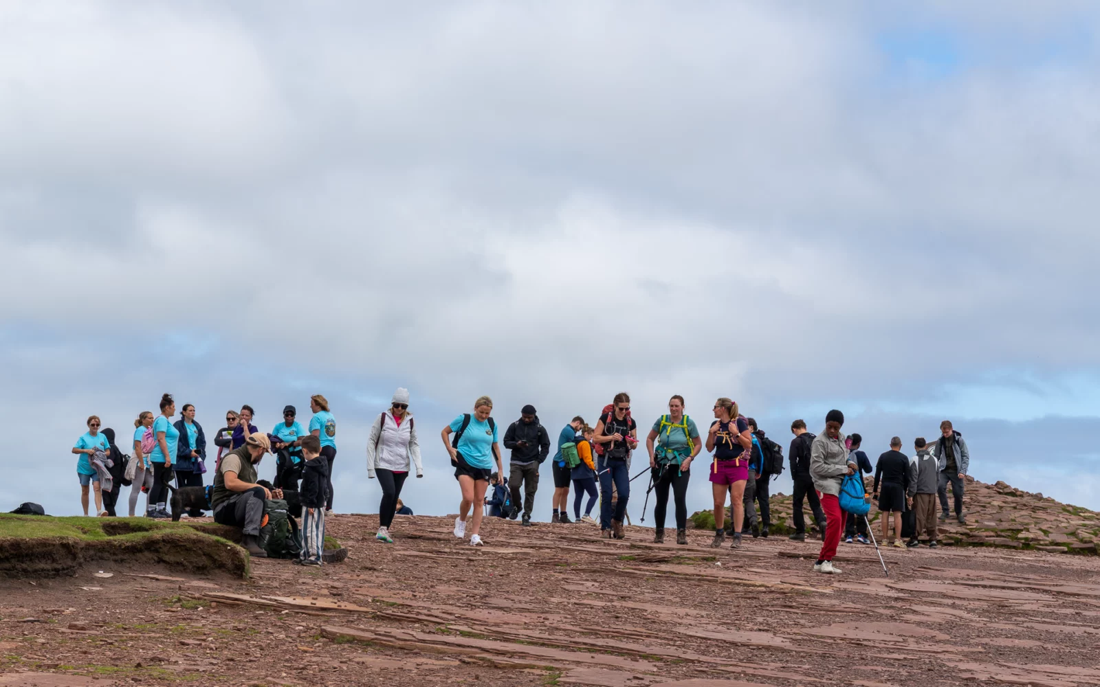 Lange rij wandelaars op de vlakke rode zandsteentop van Pen y Fan onder een bewolkte hemel, Bannau Brycheiniog in Wales