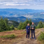 Twee wandelaars met stokken op een open bergrug bij de hut Perelik, met uitzicht over de beboste Rodopen bij Smoljan